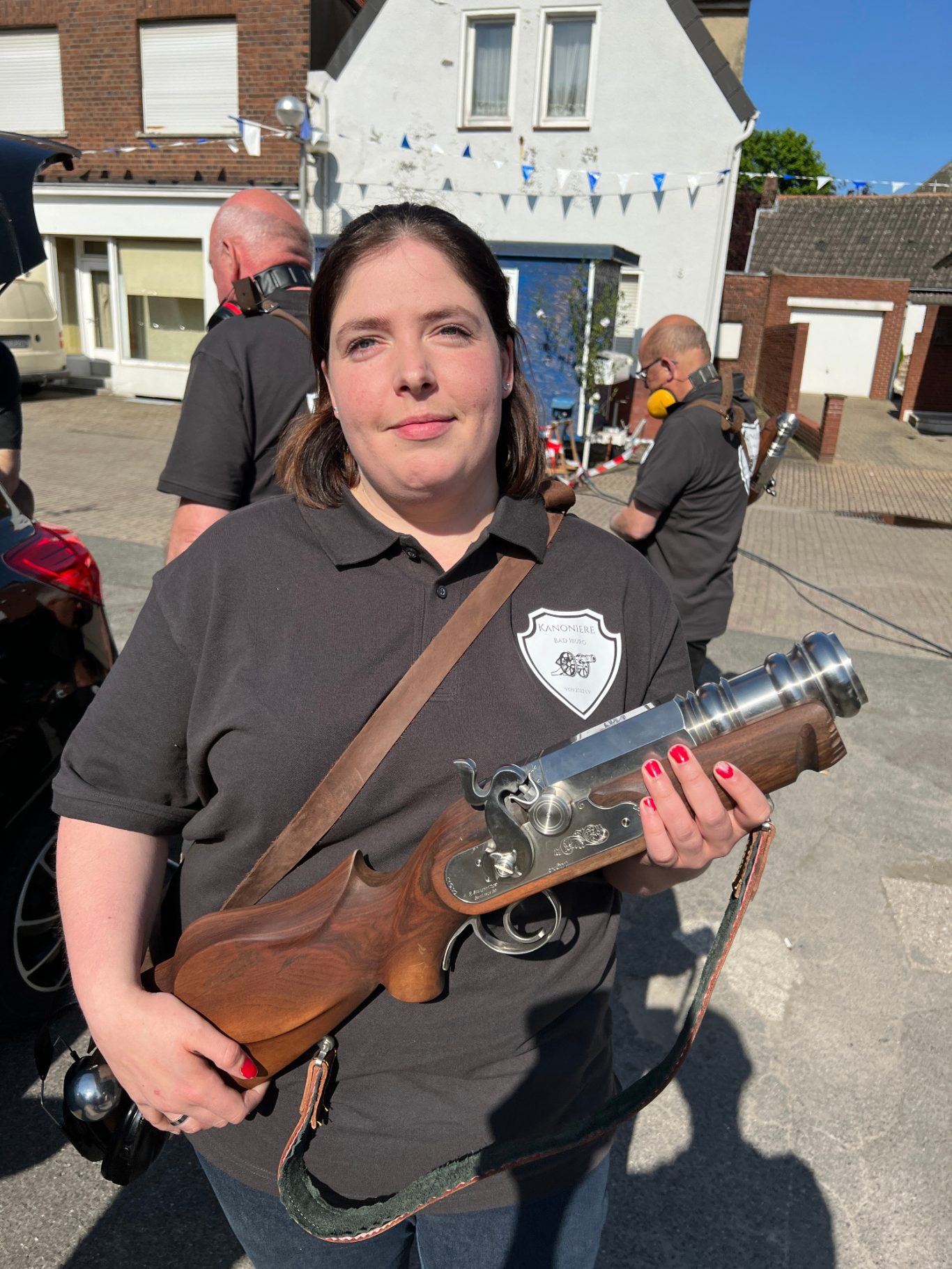 Catharina Motzkus Frau hält eine Waffe in der Hand, steht vor einem Haus mit Menschen im Hintergrund.