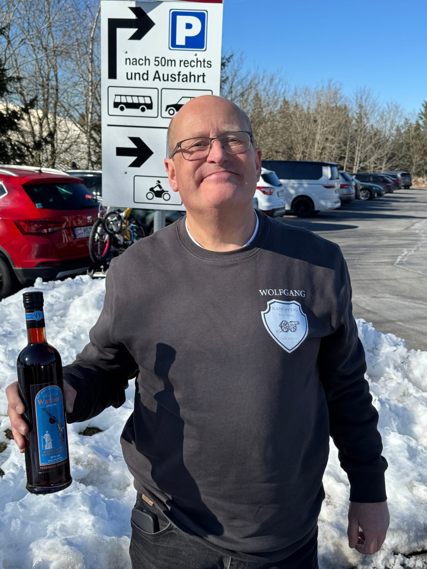 Wolfgang Müller Mann hält eine Flasche in der Hand und lächelt vor einem Parkplatzeingang.