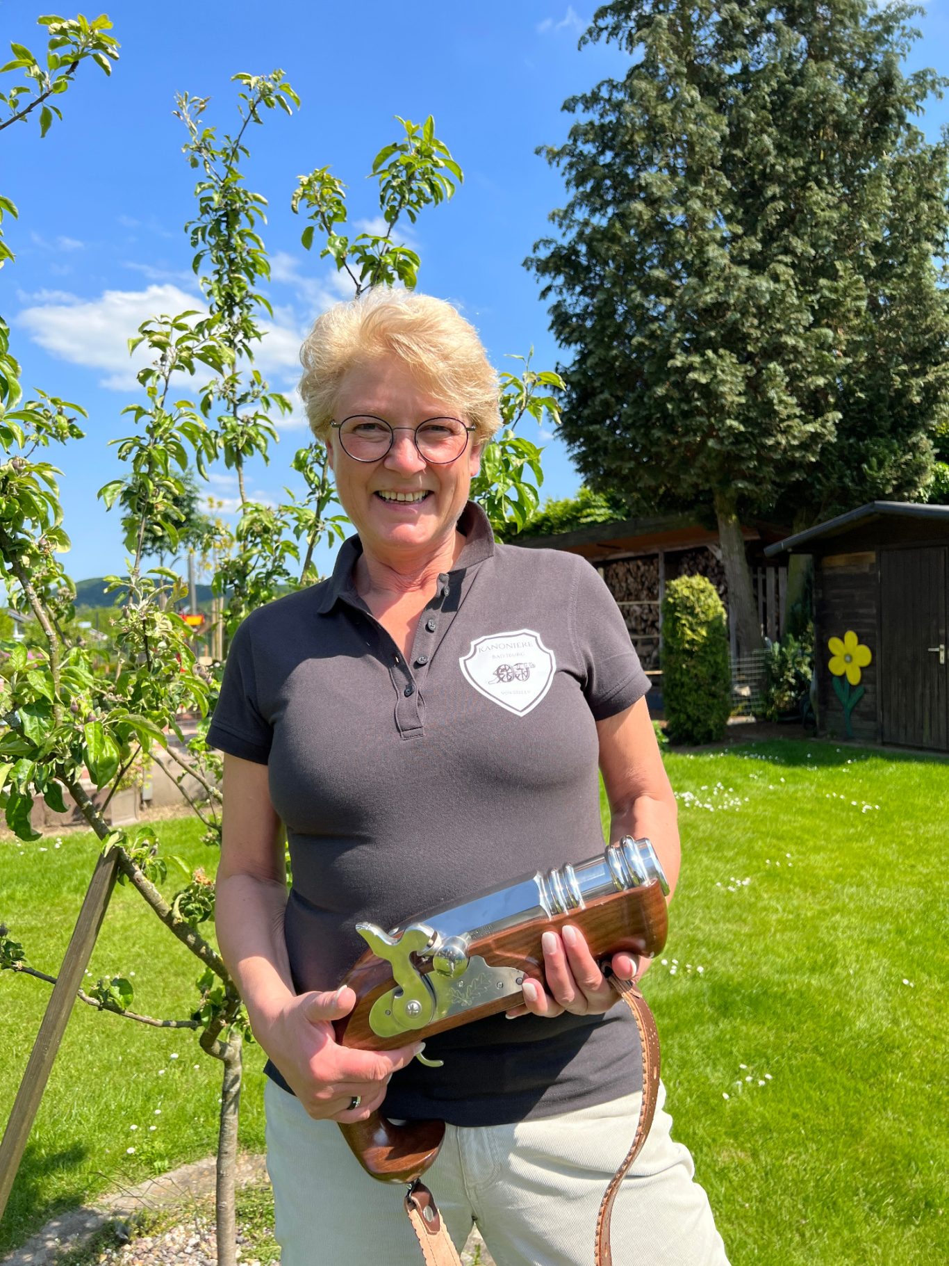 Marai Karpa Frau mit Haarschnitt, die in einem Garten eine Spritze hält, vor grünen Bäumen.