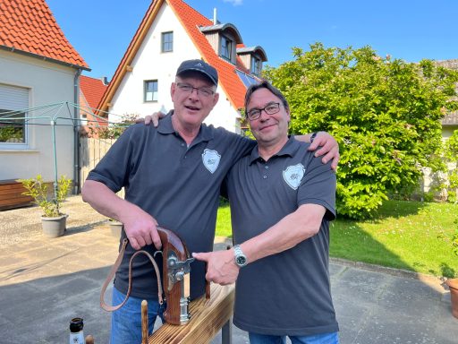 Zwei Männer in grauen T-Shirts, lächelnd und zeigend auf ein Gerät im Freien.